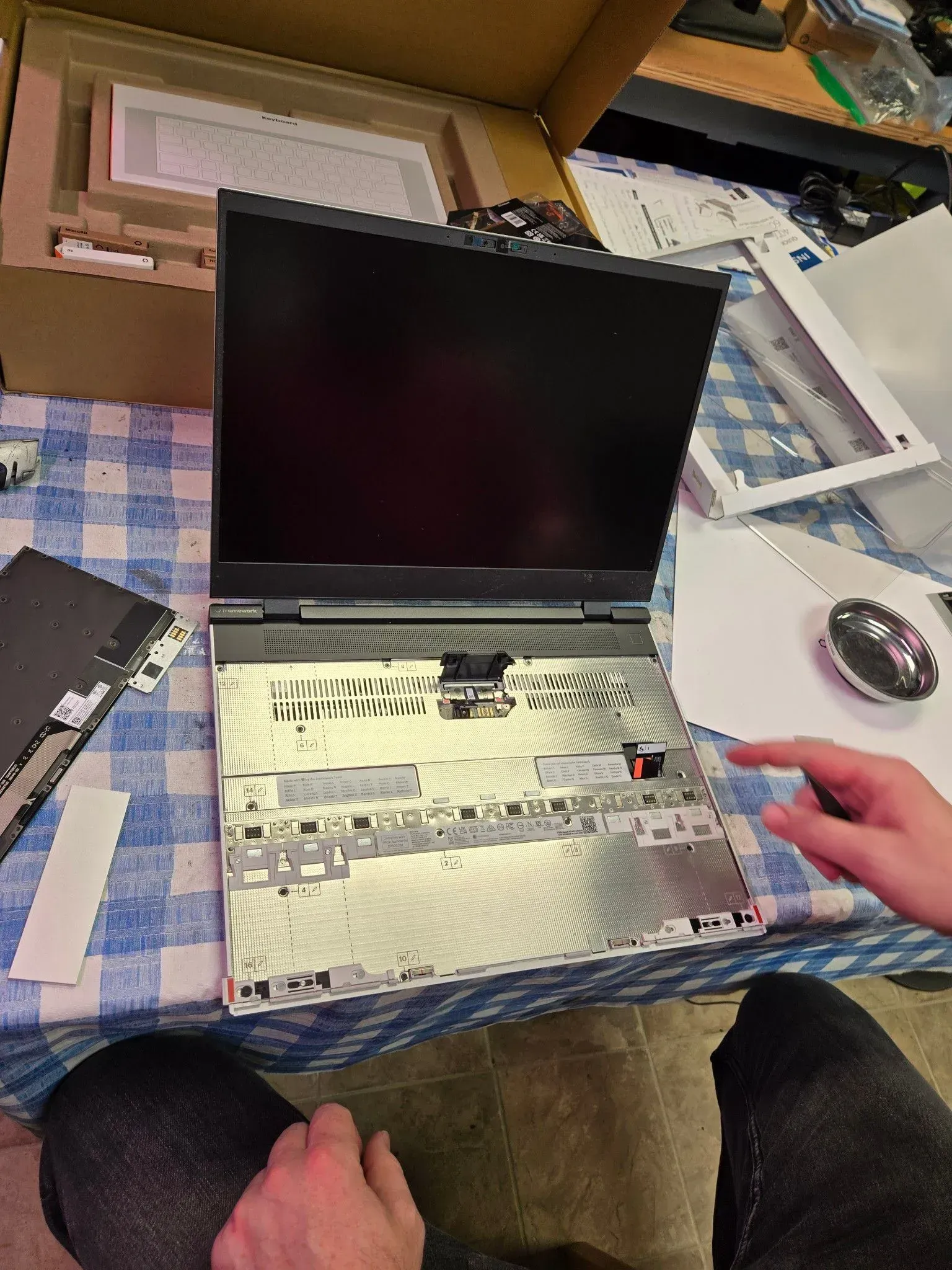 Laptop disassembled on a checkered surface, a hand points to a component. Cardboard box and tools are visible nearby.