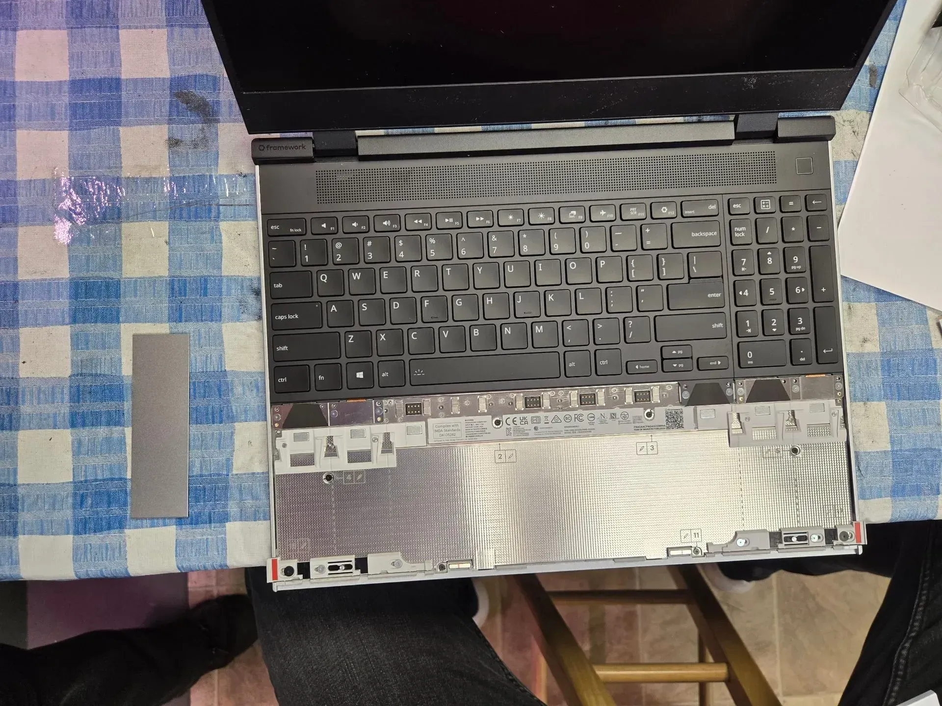 Laptop with keys removed, showing internal components. Silver metal plate next to it on a checkered tablecloth.