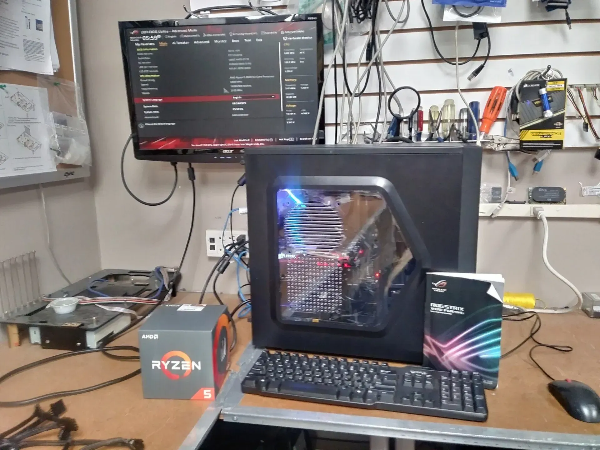 Computer tower, monitor displaying BIOS setup, Ryzen 5 box, keyboard, mouse on a desk.