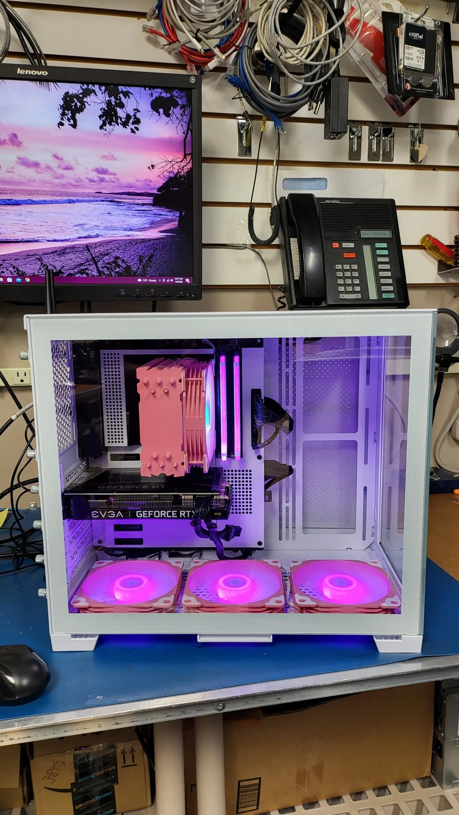 White computer case with pink and purple lighting, set on a blue surface, with a monitor and phone in the background.