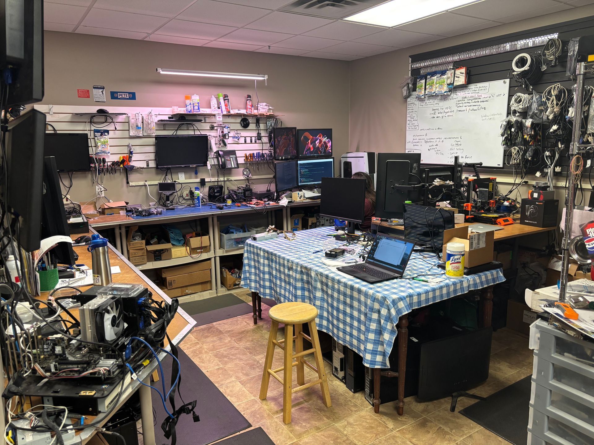 A cluttered tech repair workshop with computers, tools, and a workbench covered in wires and screens.
