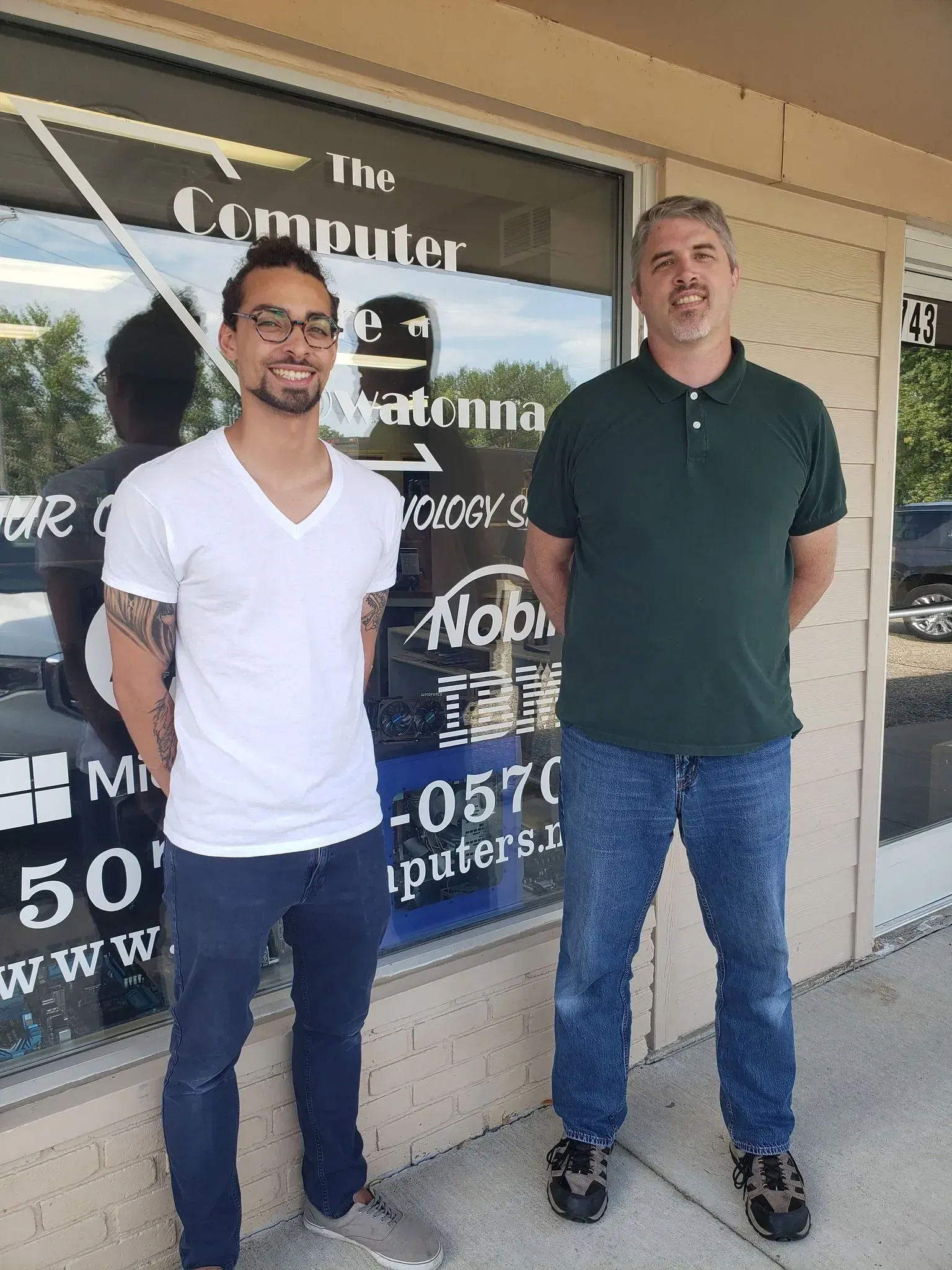 Two men stand in front of a computer repair shop. One wears a white shirt, jeans. The other wears green polo and jeans.