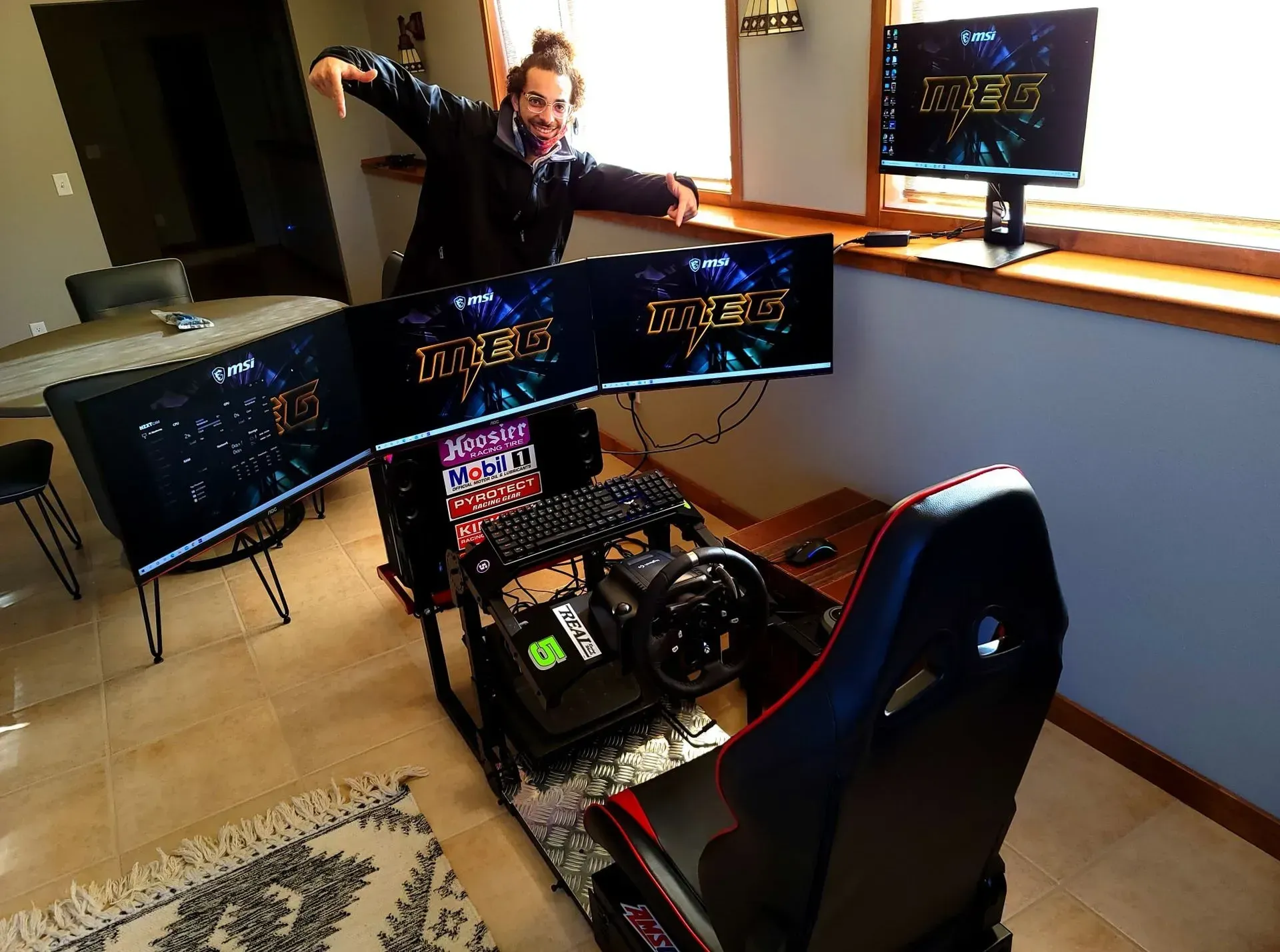Man points at triple-screen racing simulator setup in a room with a window, displaying a game logo.