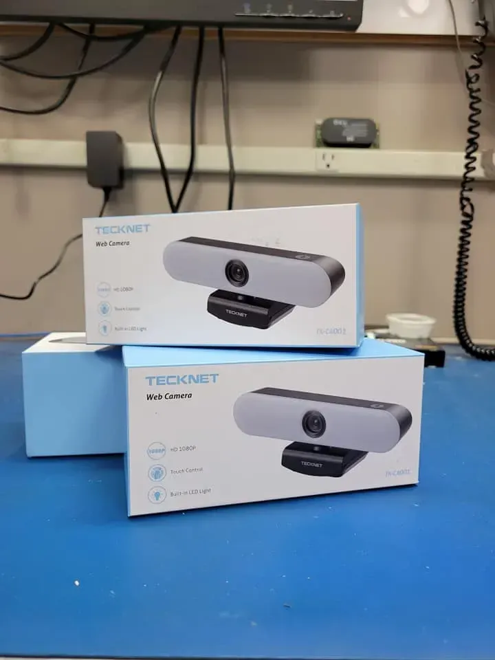 Two TeckNet webcams in boxes stacked on a blue surface, against a background of electronics.