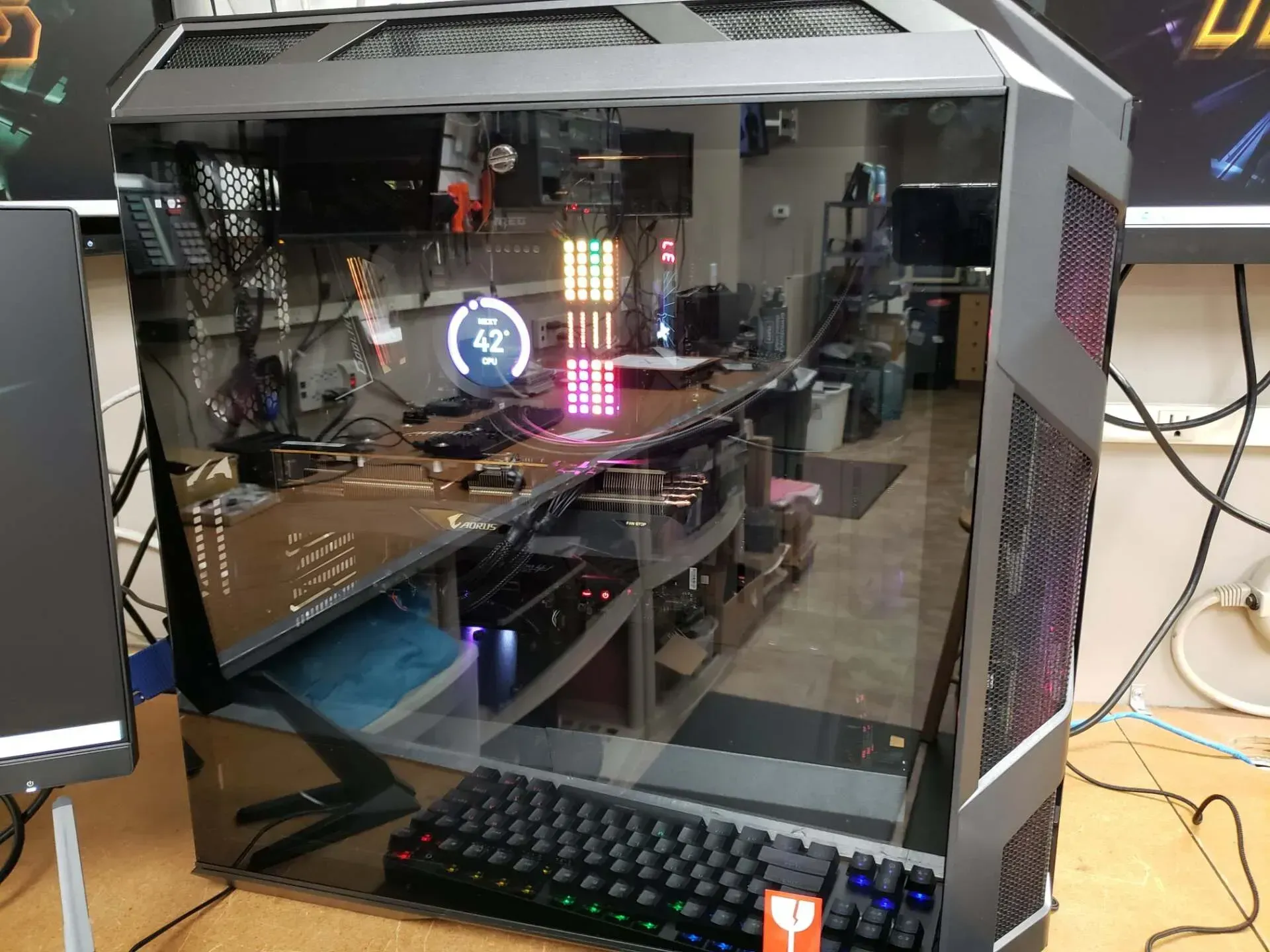 Large, clear PC tower case with a keyboard in front, reflecting an office setting with computer parts and equipment.