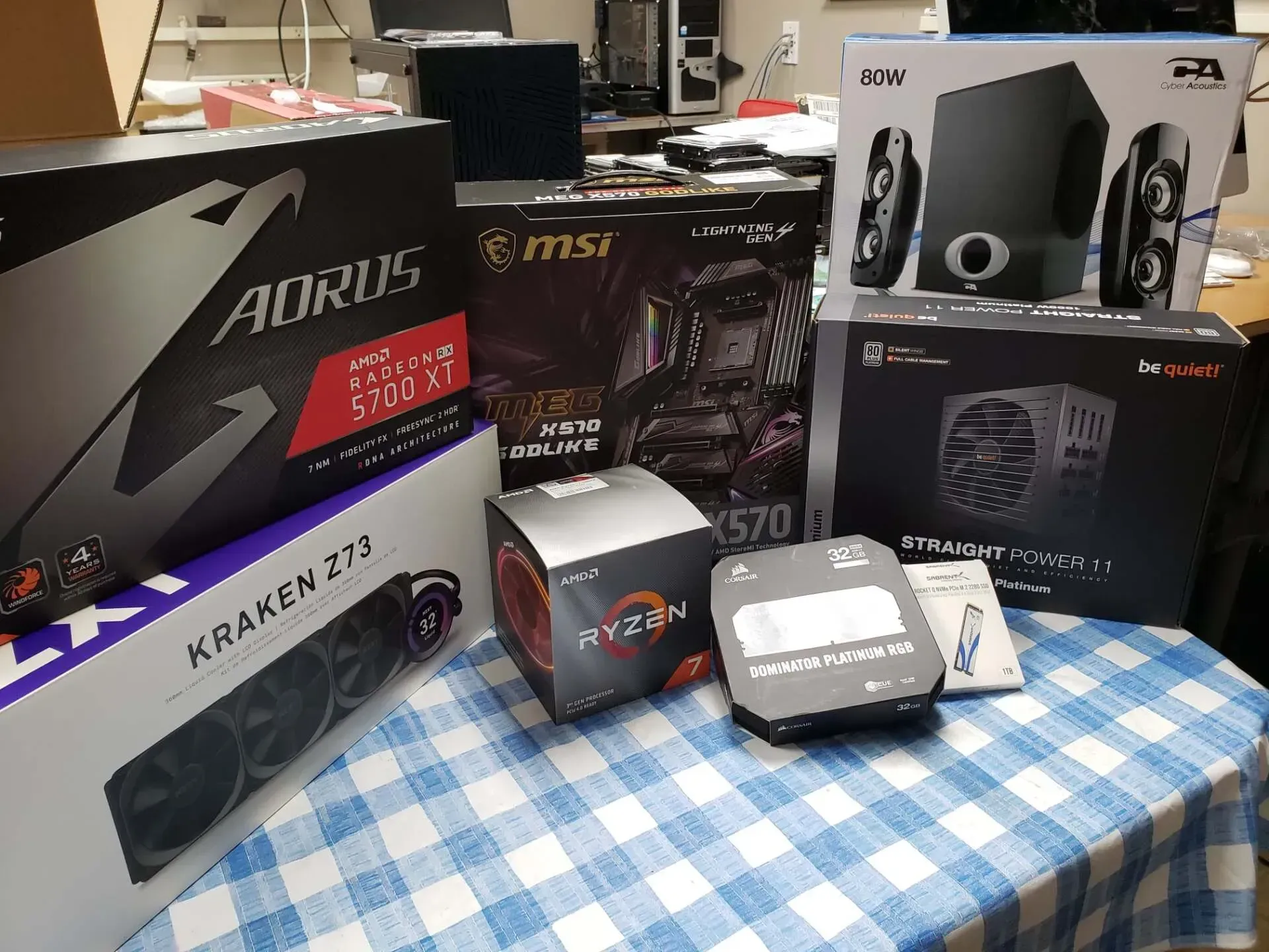 Computer components boxes on a checkered tablecloth: GPU, CPU, motherboard, cooler, speakers, SSD.