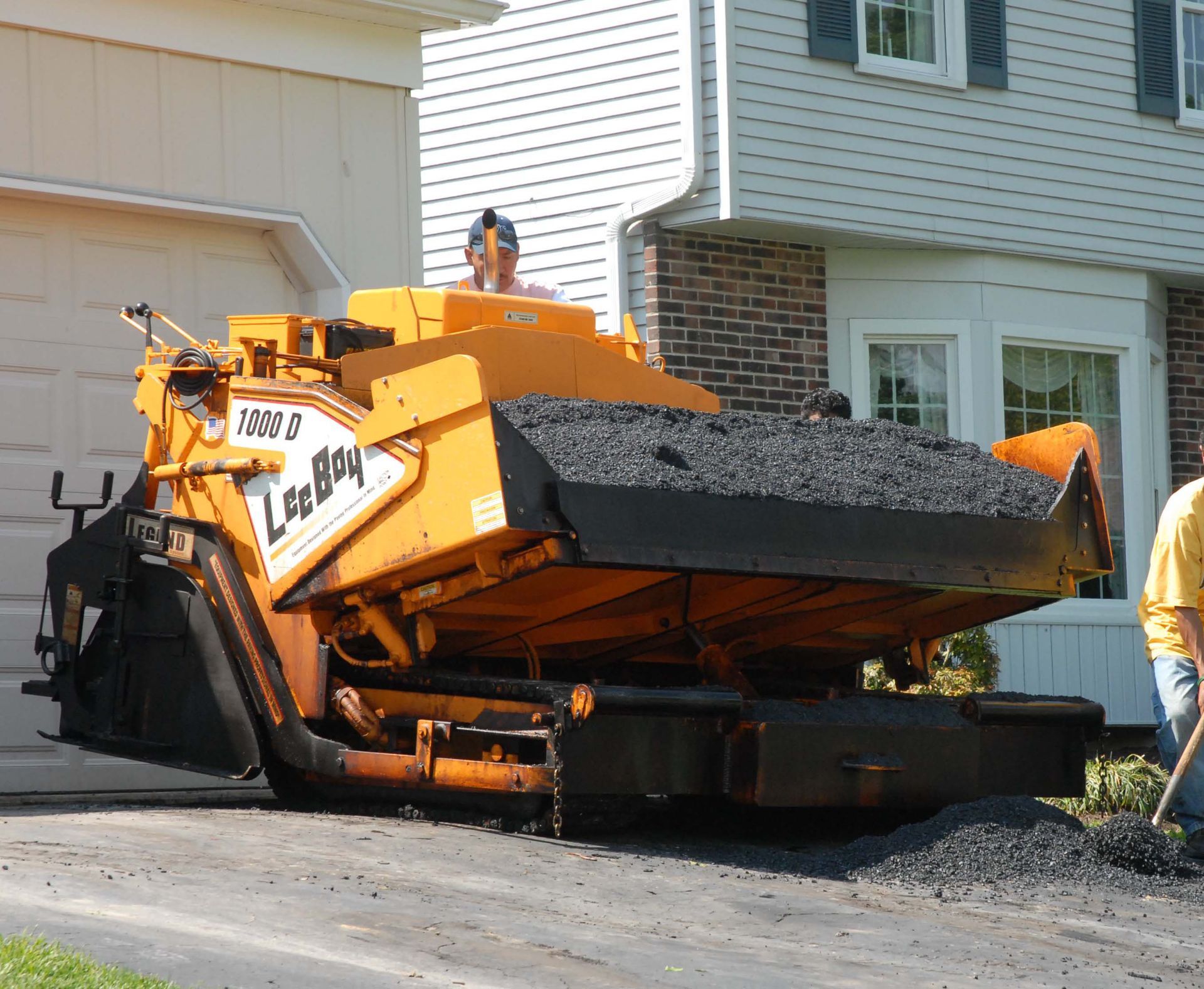 A yellow LeeBoy asphalt paver spreading material on a residential driveway in front of a house.