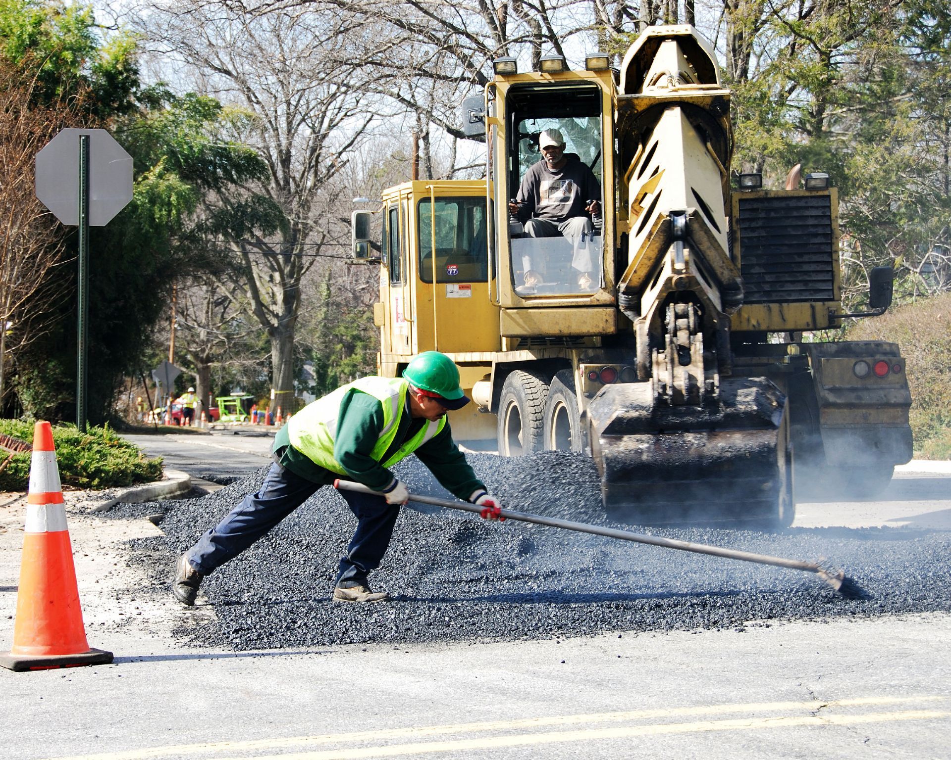 A worker in a high-visibility vest spreads hot asphalt on a road near a yellow construction vehicle and a safety cone.