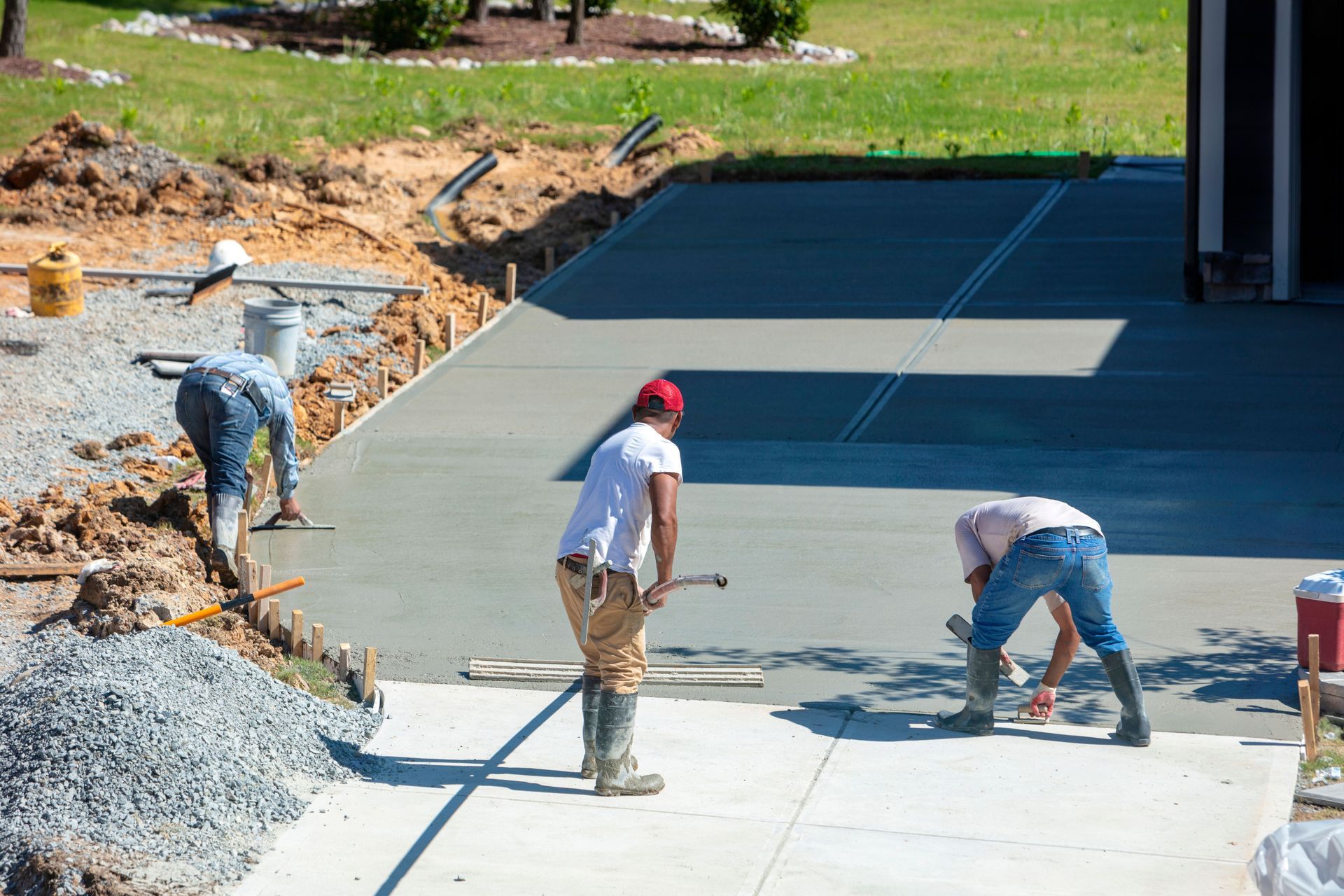 Three construction workers finish smoothing a freshly poured concrete driveway in a sunny residential yard.