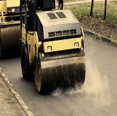 A yellow steamroller compacts fresh asphalt on a road, creating a cloud of dust.