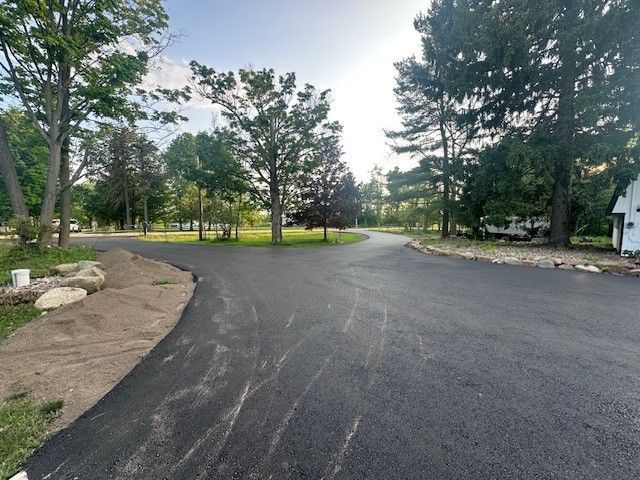 A worker in high-visibility orange and navy gear uses a shovel to smooth fresh black asphalt on a road near two rollers.