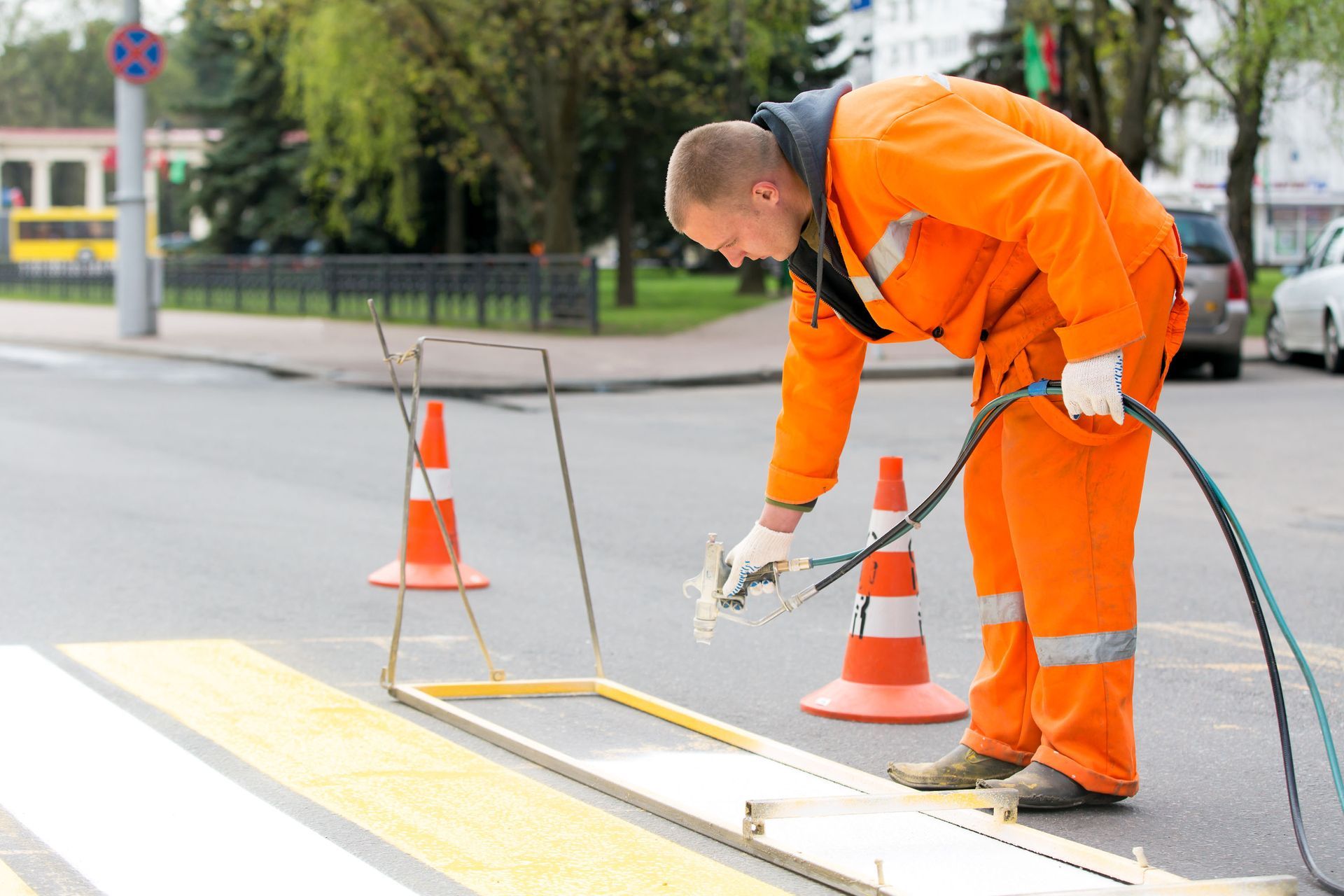 A worker in bright orange coveralls uses a spray device to paint white stripes on a crosswalk with the aid of a stencil.