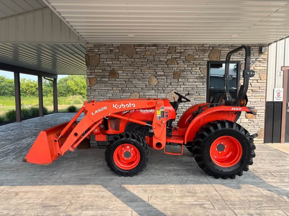 Orange Kubota tractor with a front loader parked outdoors near a stone wall.