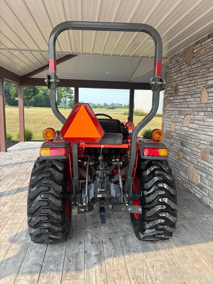 Rear view of an orange tractor parked under a covered area with an orange safety triangle, taillights, and a rollover protection system.