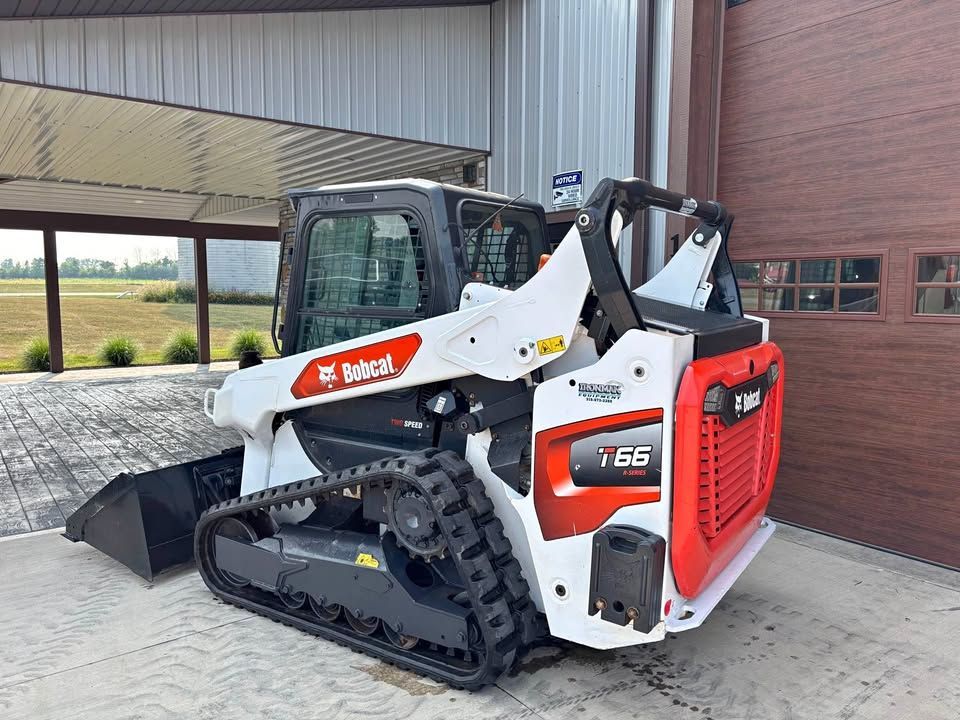 A white and red Bobcat T66 compact track loader parked near a building with a loading bucket.