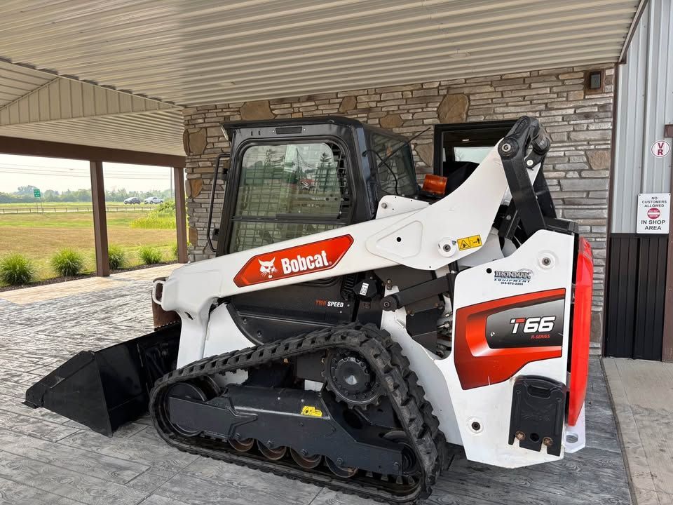 White and red Bobcat compact track loader parked under a canopy. The loader is on a concrete surface with a field in the background.