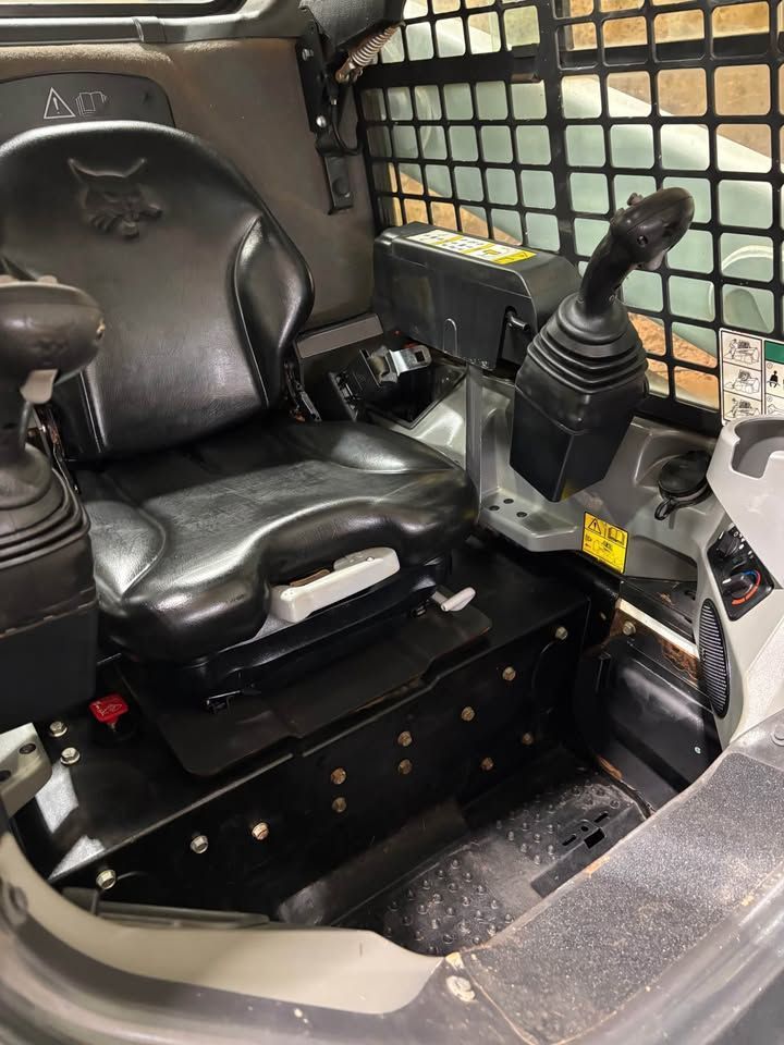 Interior of a Bobcat skid steer. Black seat and joystick controls, safety bars, and metal floor.
