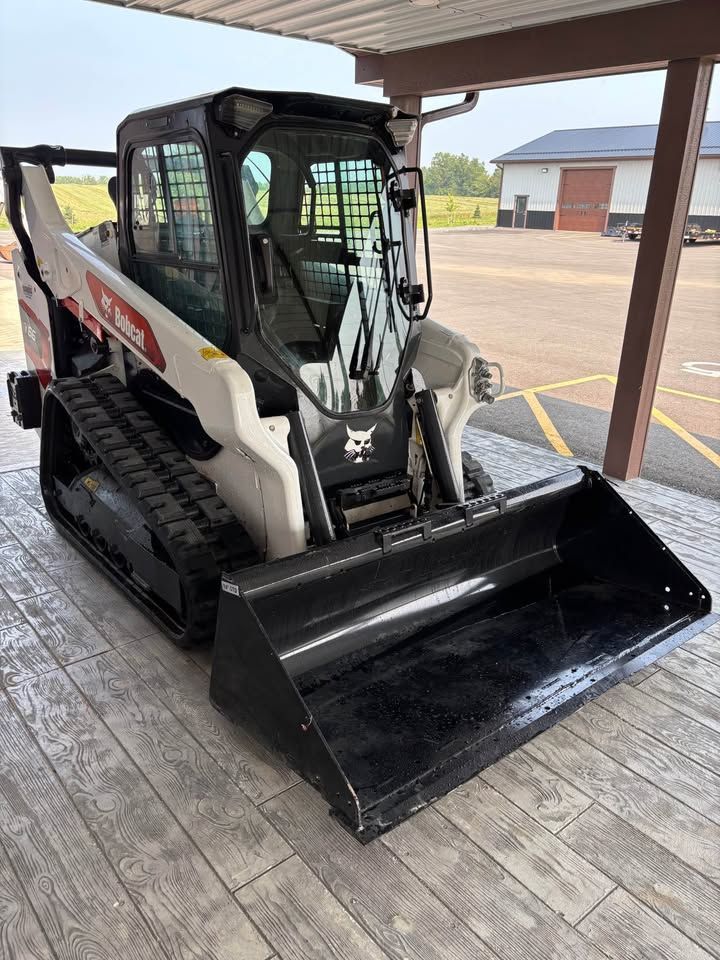 White Bobcat track loader with a black bucket parked on a gray surface under a covered area. A building is visible in the background.