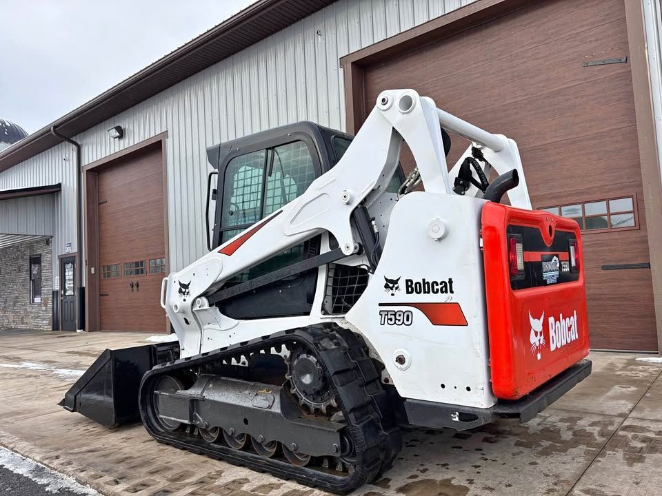 White and orange Bobcat T590 track skid steer loader in front of a building with brown garage doors.