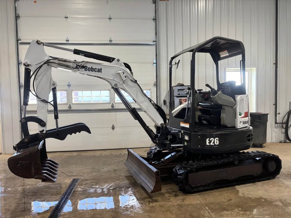 Bobcat E26 mini excavator in a garage. White and black, with a bucket and tracks.