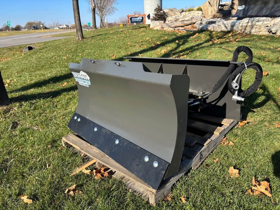 An olive-green snow plow on a wooden pallet sits on grass near a road on a sunny day.