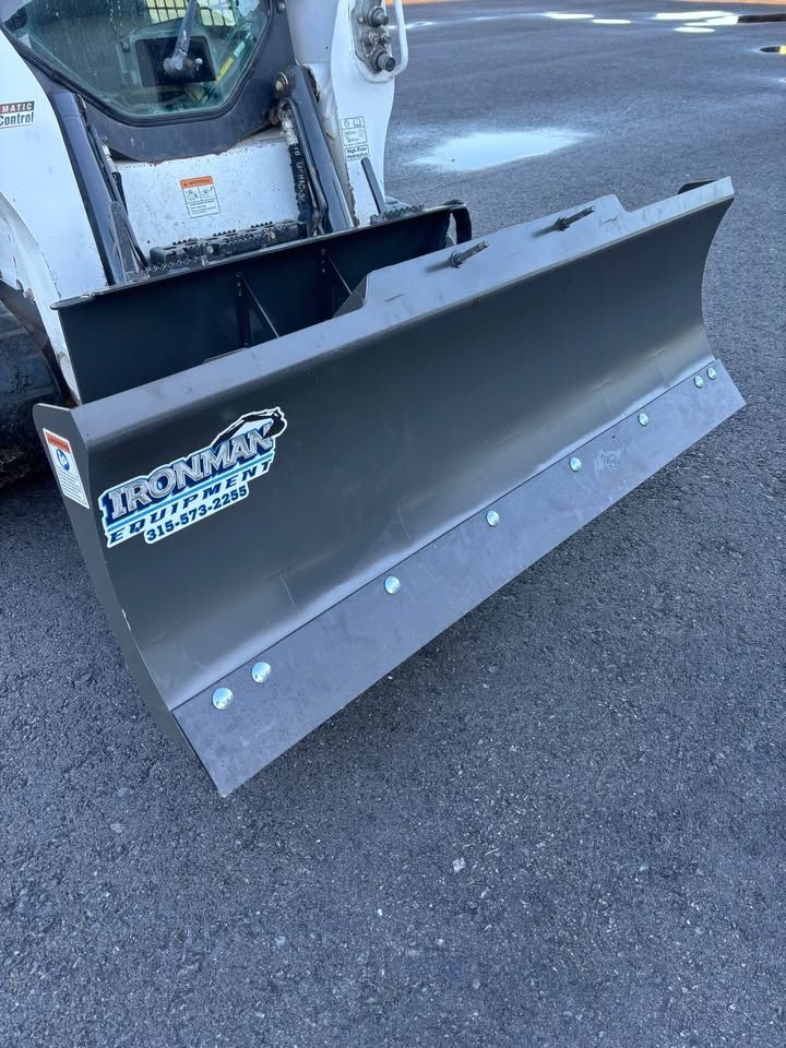 Black snowplow attached to a Bobcat machine, angled slightly down, on pavement.