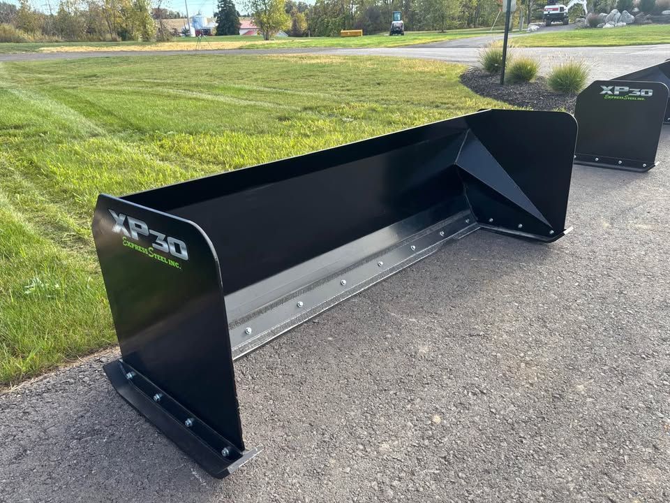 Black XP30 snowplow blade on a paved surface, outside. Grass and sky in the background.