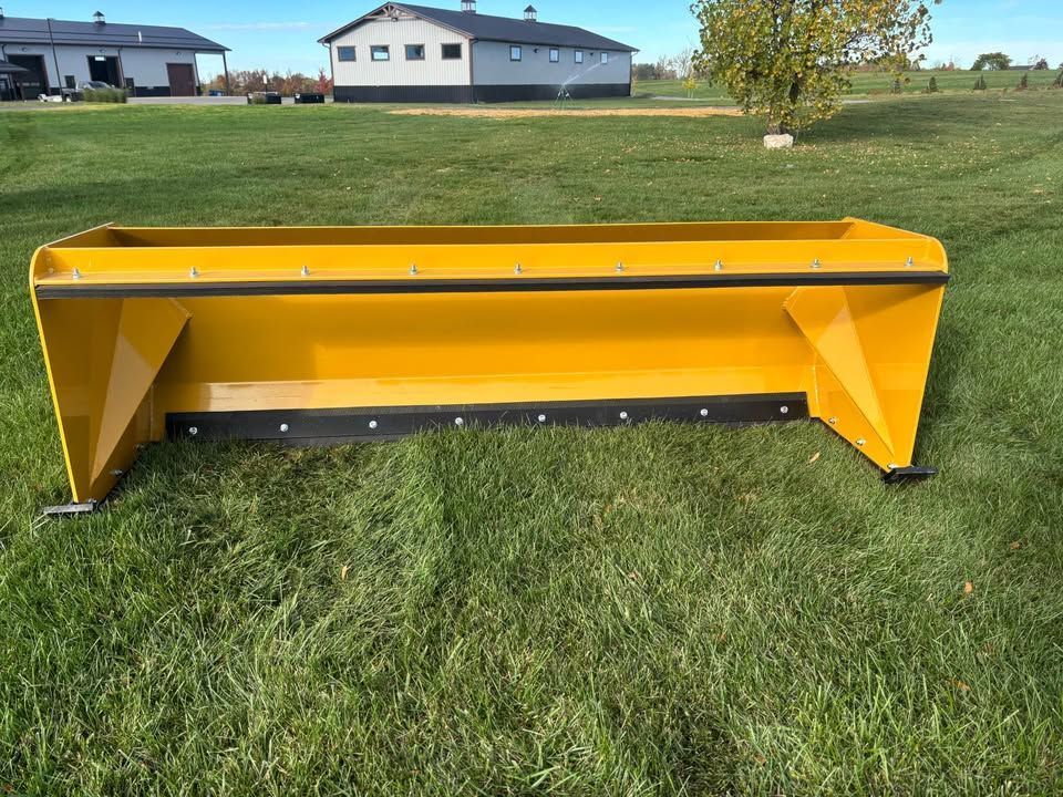 Yellow snowplow blade on green grass, in front of buildings.