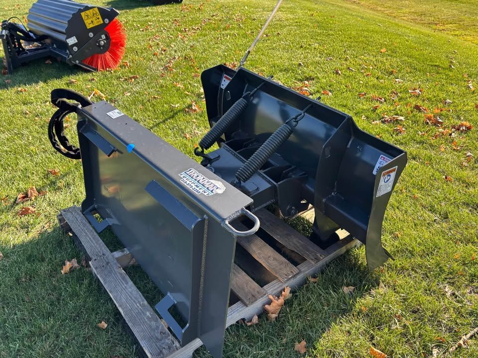 Dark gray snowplow attachment on a pallet outdoors. Brush attachment visible in the background.