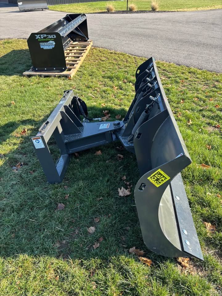 Two snowplow attachments on grass. The larger one is angled and gray with a yellow sticker.