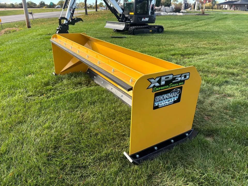 Yellow snow pusher attachment on grass, with an excavator in the background.