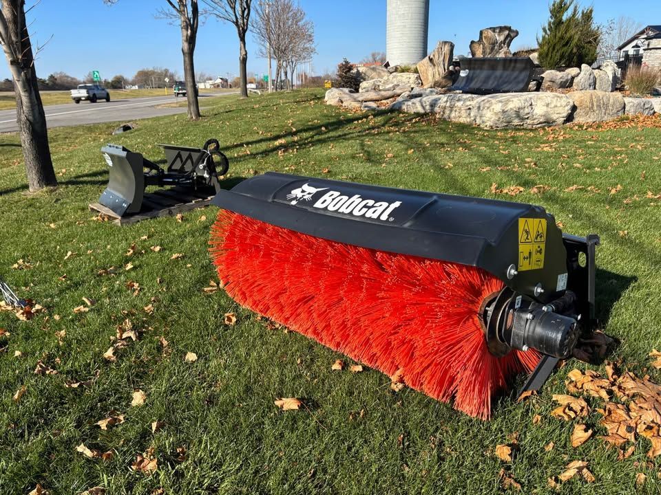Bobcat rotary broom on grass, clearing leaves. Black with red bristles, outdoors.