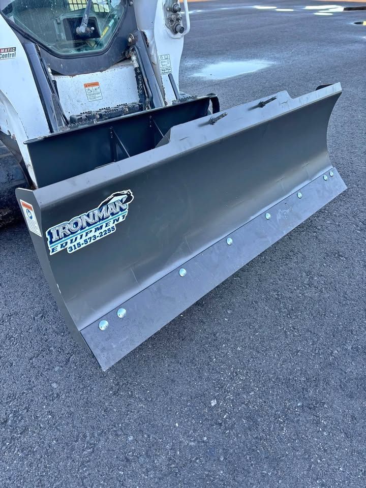 Gray snow plow attached to a white skid steer, parked on pavement.