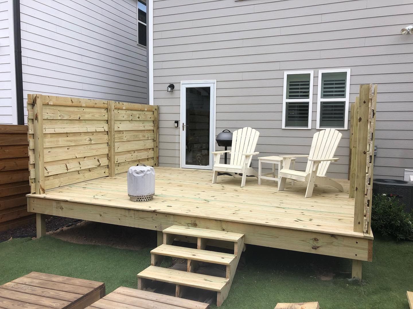 Small wooden backyard deck with steps, railing, and two white chairs beside a house wall