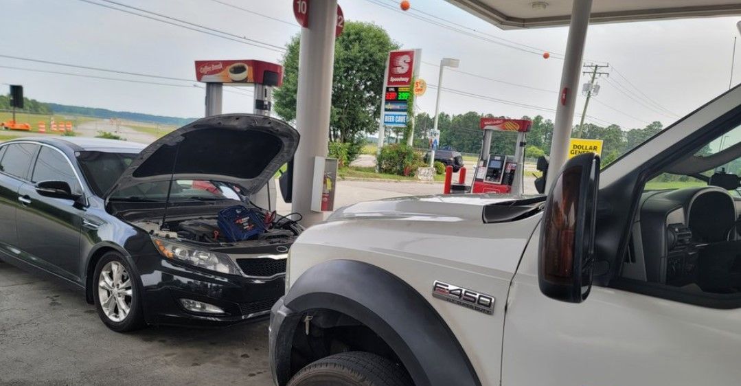 A car with its hood up is parked next to a truck at a gas station.