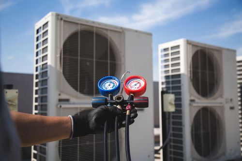Hand holding HVAC gauges in front of outdoor air conditioning units