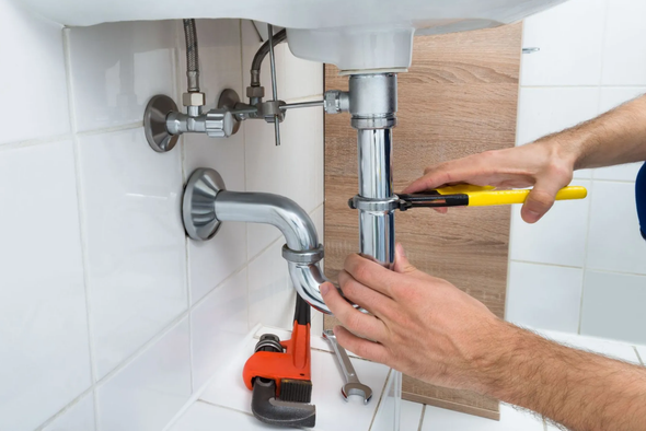A plumber uses pliers to tighten a pipe under a white sink in a tiled bathroom.