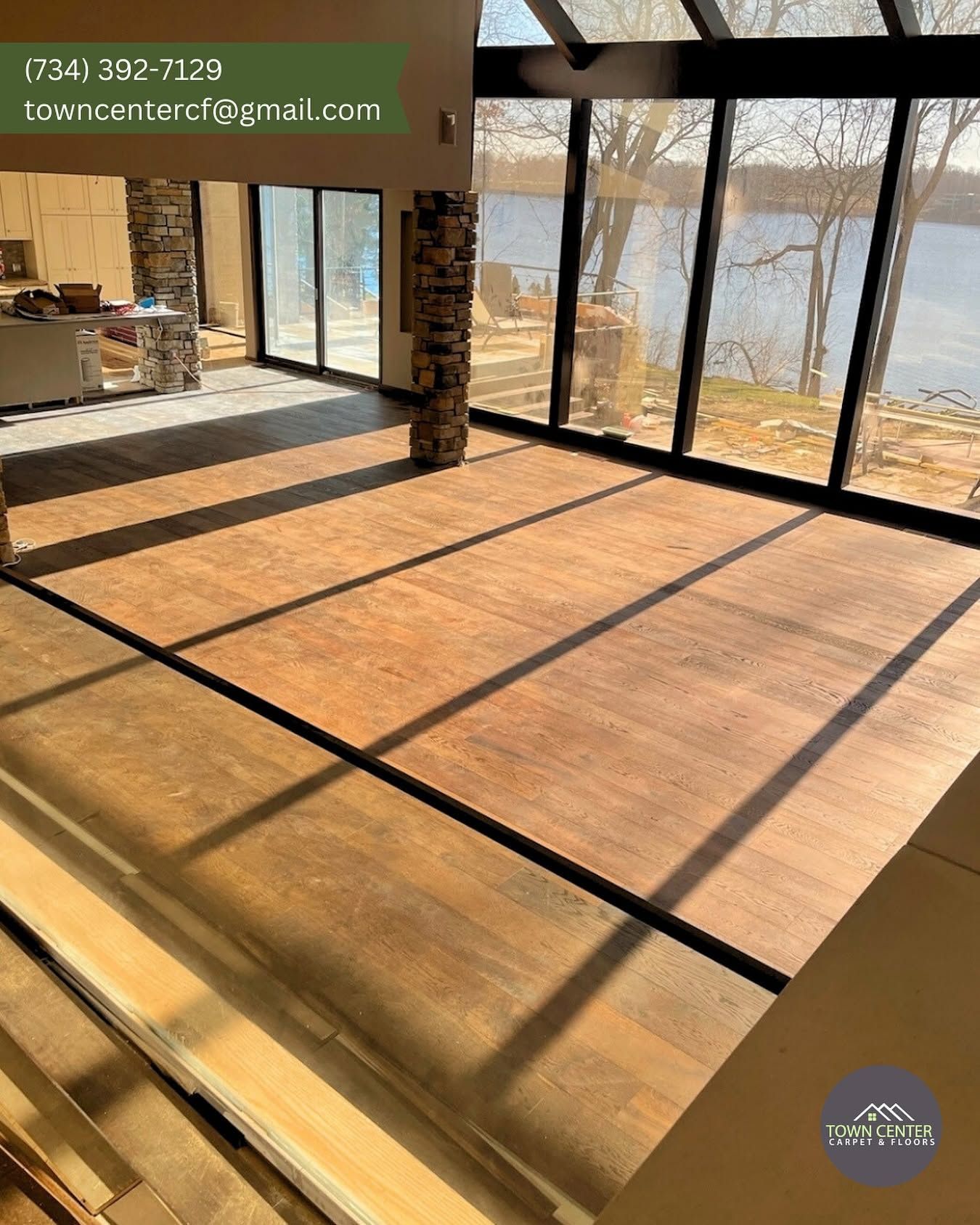 Sunlit empty room with floor-to-ceiling windows, black frames, and warm wood floors overlooking water.