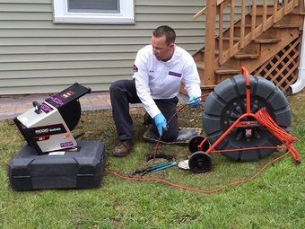 Man inspecting a sewer line with equipment on grass near a house.