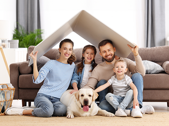 Family of four and dog under a cardboard roof, smiling in a living room.