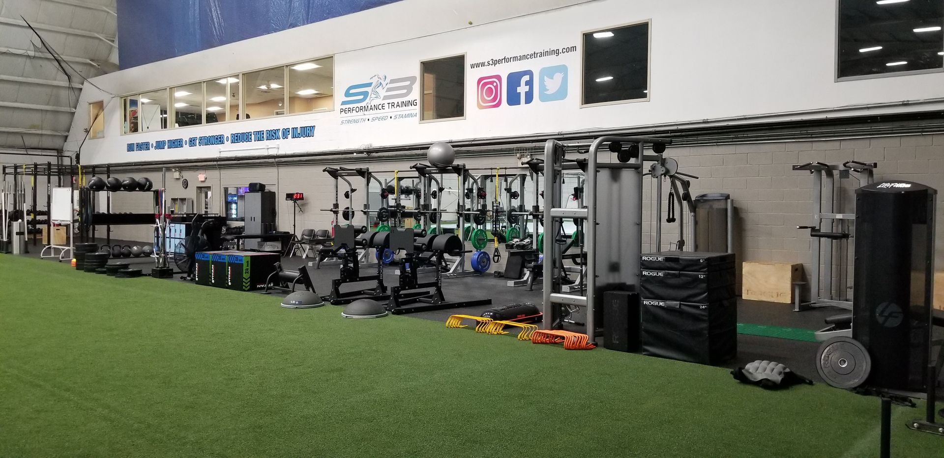 Indoor gym training area with exercise machines, free weights, and turf lane along a wall of logos and posters