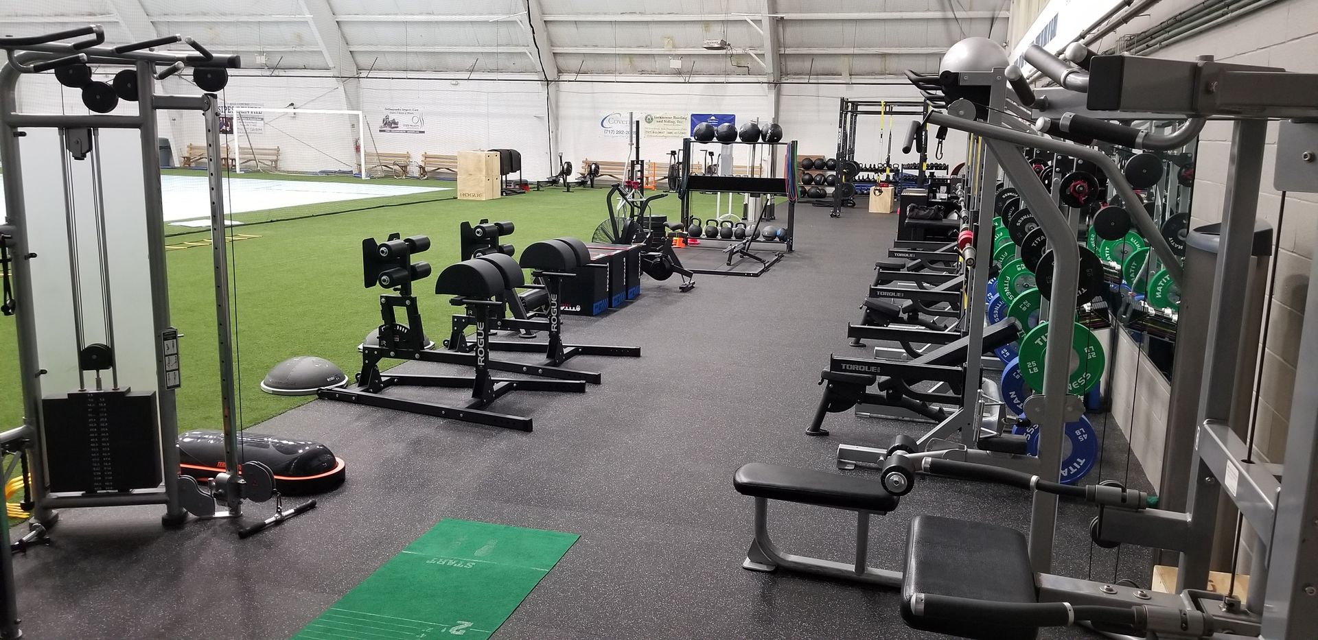 Indoor gym with weight machines, benches, and turf training area under bright lights
