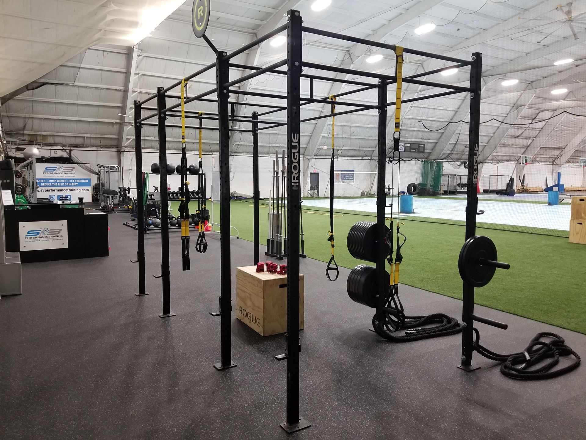Indoor gym with black squat racks and hanging straps on green turf under a white ceiling