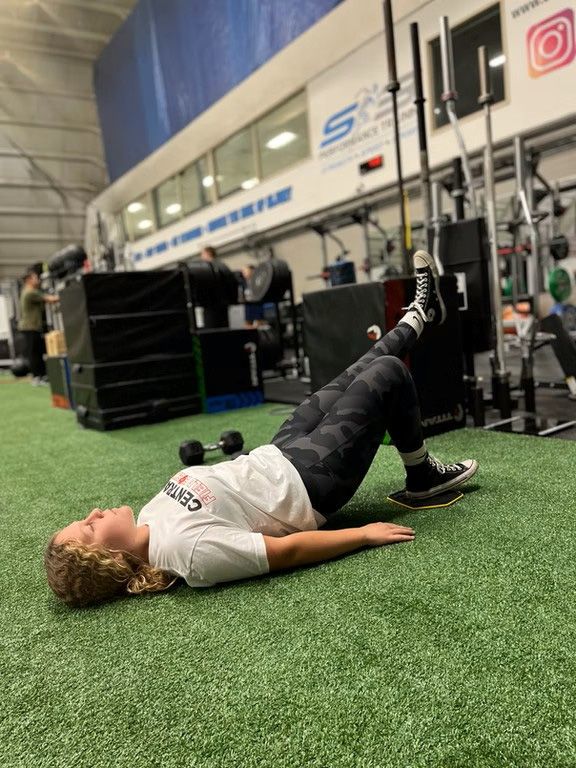 Person doing a bridge exercise on turf in a gym, with weight equipment in the background