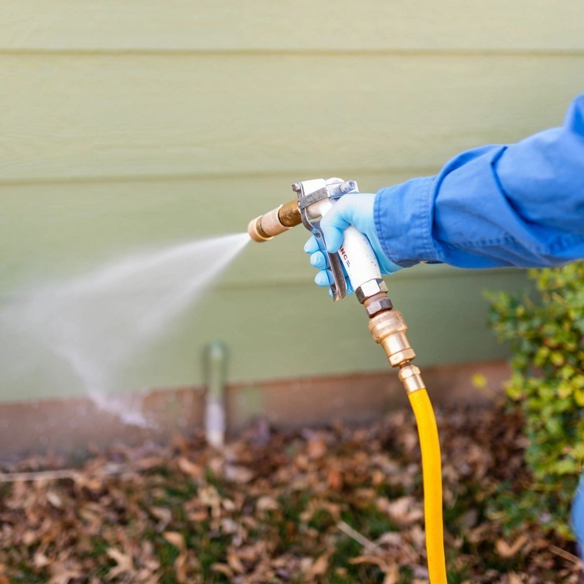 A person wearing a blue shirt and protective glove uses a yellow hose with a spray nozzle to water a light green wall.
