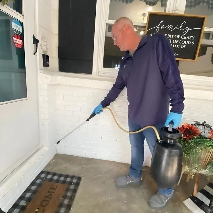 A person in a blue long-sleeved shirt and gloves sprays the perimeter of a white doorway with a handheld pump sprayer.