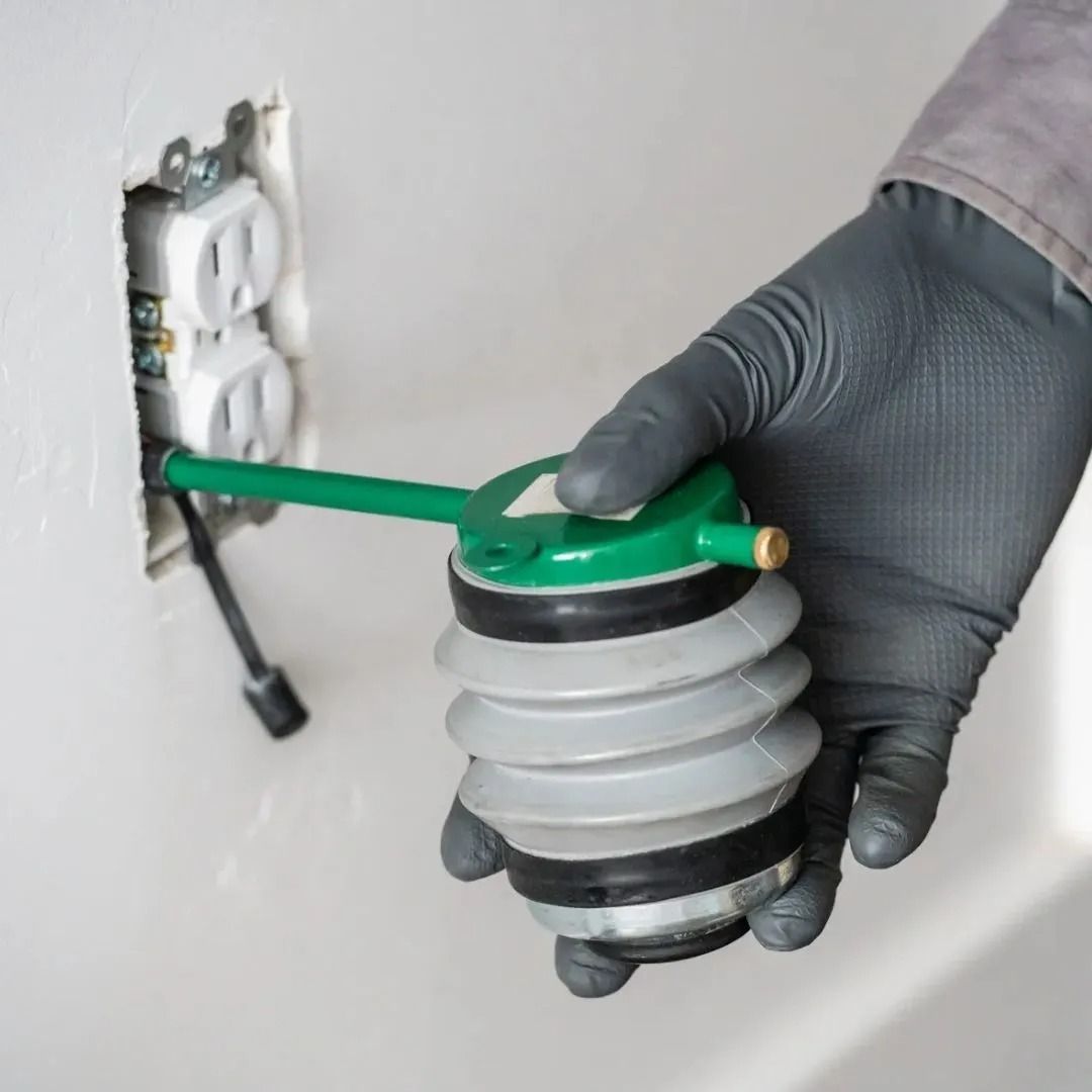 A gloved hand uses a green-and-gray bellows-style powder duster to apply pest control dust into an electrical outlet.