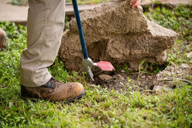 A person in work boots uses a tool to lever a large, light-brown rock out of the grass.