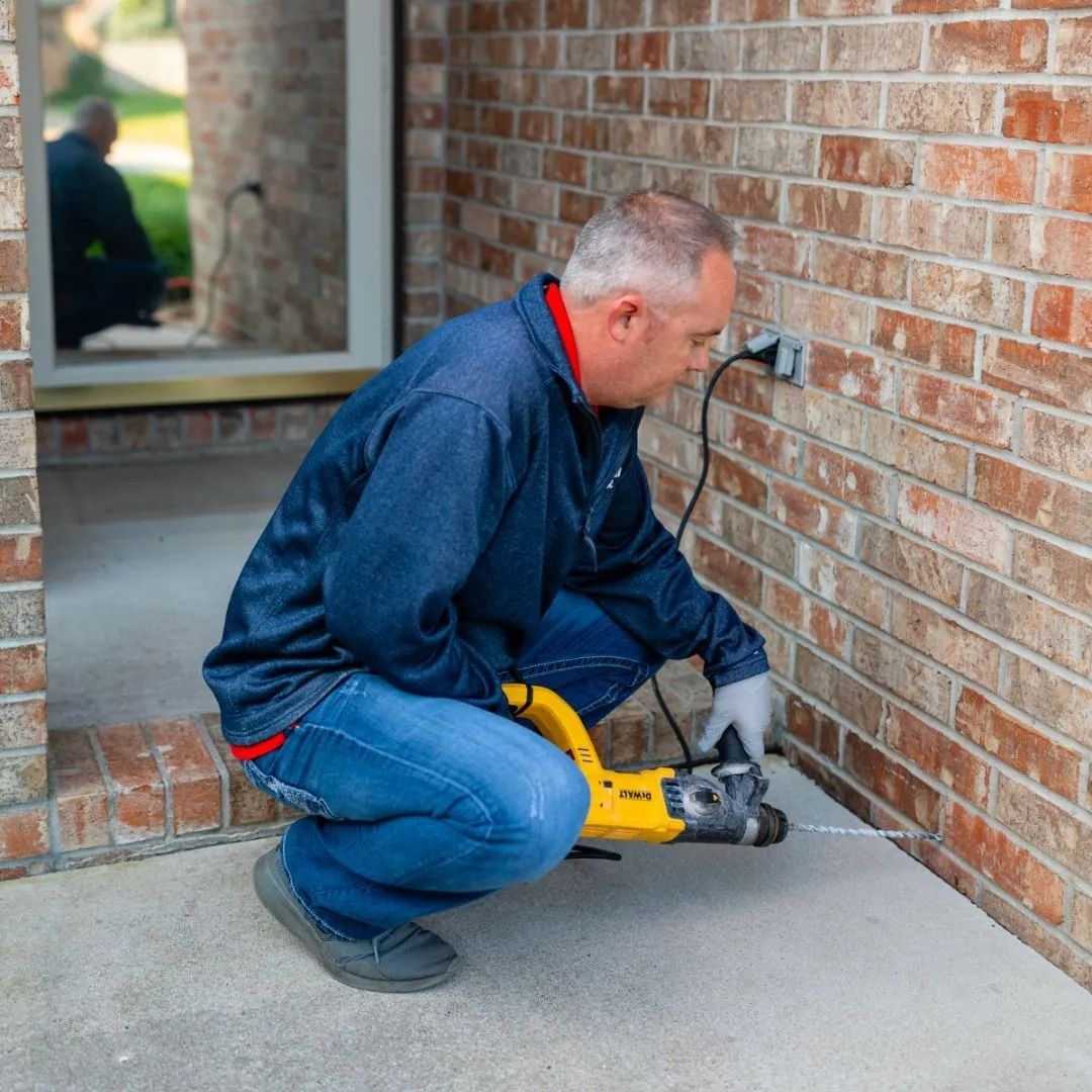 A person kneels on a concrete porch, using a yellow power drill to create a hole in the base of a brick exterior wall.