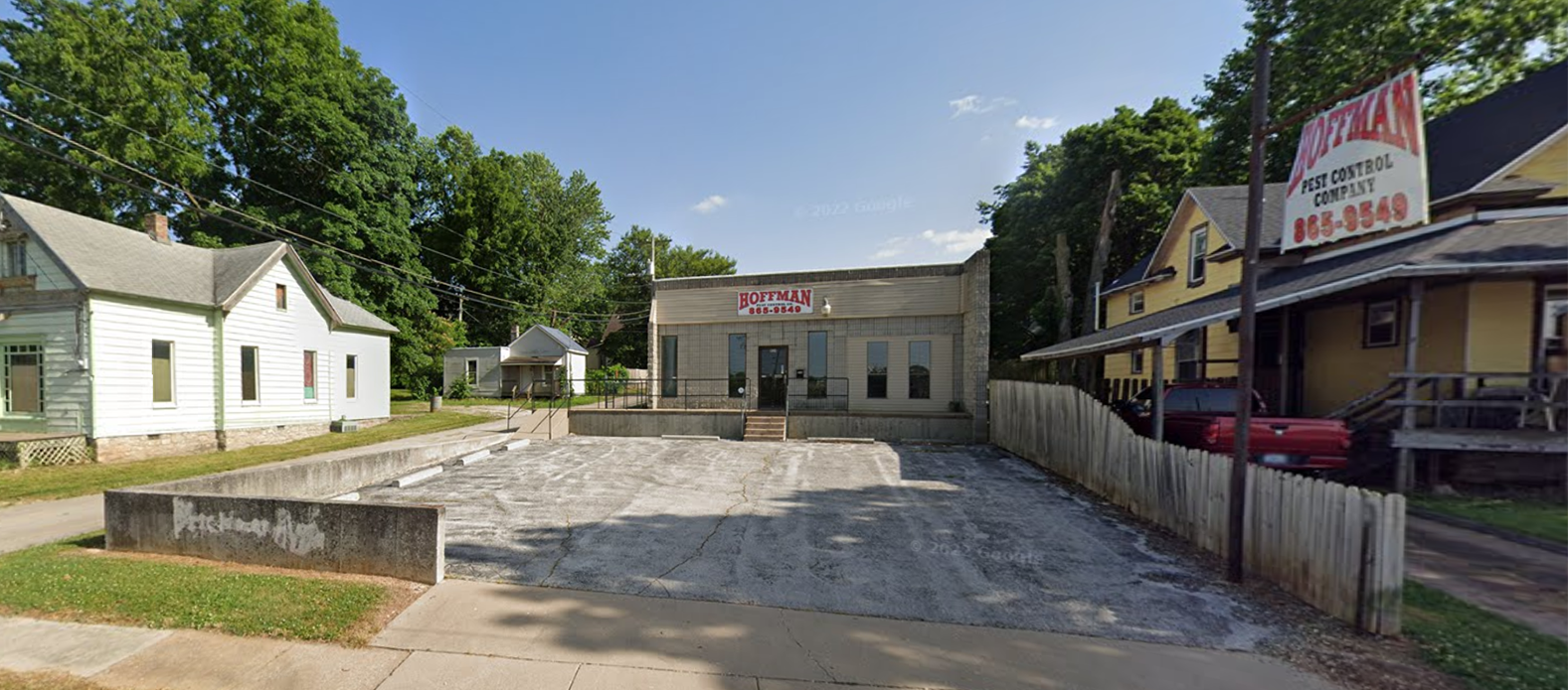 A small, beige storefront sits behind a gravel lot in a rural town, flanked by white and yellow houses under a sunny sky.